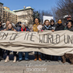 At the NYC Women’s March 2025 in Union Square, New York City, activists Leslie Cagan, Yanery Cruz, Evie K, Kim Russell, and Evie Joy stand united, holding a large fabric banner reading "WOMEN OF THE WORLD UNITE." The powerful message stretches across the group, symbolizing global solidarity in the fight for women’s rights. The diverse participants, dressed in warm clothing, represent a mix of seasoned activists and younger voices. Behind them, other demonstrators hold signs advocating for feminist and social justice causes, while some wear sunglasses, hats, and scarves against the chilly air. A brown dog sits at the feet of one of the participants, adding a community-driven touch to the gathering. The atmosphere is one of determination, unity, and activism, as the women stand together for gender equality, reproductive rights, and justice.