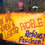 At Union Square in New York City, a group of activists at the NYC Women’s March 2025 hold a bold, spray-painted banner reading "STOP THE WAR ON WOMEN & LGBTQ PEOPLE! REFUSE FASCISM!" The vibrant yellow and red sign stands out among the crowd, amplifying the urgency of the message. The woman in the center wears a pink pussy hat, a symbol of feminist resistance, and a thick scarf wrapped around her face against the cold. To her left, another protester wears a knit orange beanie and sunglasses, adding to the atmosphere of defiance and determination. The background features other demonstrators holding signs and banners, with trees and city buildings framing the scene. The energy in the gathering is palpable as marchers call for gender equality, LGBTQ rights, and opposition to oppressive policies.