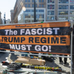 A large black and orange banner with bold white text reading "The FASCIST TRUMP REGIME MUST GO!" is prominently displayed at Union Square during the NYC Women’s March 2025. The banner is supported by a metal frame and includes the website "RefuseFascism.org," an organization known for its advocacy against authoritarianism and far-right politics. A woman in a black winter coat, sunglasses, and knit gloves stands beside the sign, looking towards the camera with a composed expression. In the background, a man in a black beanie and sunglasses holds up a phone, possibly taking a photo or recording the event. Stacks of protest signs, banners, and poles are laid on the ground, suggesting active participation in the demonstration. The event takes place in the bustling public space of Union Square, with recognizable storefronts, including Burlington and Sephora, visible in the background. The scene captures the political resistance, activism, and energy of the protest movement.