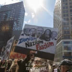 A group of protesters participates in the Women's March on March 8, 2025, from Washington Square Park to Union Square, raising signs expressing their political views. In the foreground, an individual wearing a knitted beanie, sunglasses, and a colorful scarf holds up a large sign with bold text reading: "Zionism = Nazism = Fascism." The sign features edited images of several political figures, including former U.S. President Donald Trump, Israeli Prime Minister Benjamin Netanyahu, U.S. Vice President Kamala Harris, and U.S. President Joe Biden. The demonstrator's hand is visible, gripping the wooden stick supporting the sign.
Surrounding this protester, other individuals are also holding up signs, including one with the words "NOW" and another featuring a heart symbol. The setting is Union Square, with tall buildings, a clock mural, and a clear blue sky in the background. The sunlight creates a striking contrast between the demonstrators and the cityscape. The march is part of a global movement advocating for women's rights, social justice, and political accountability.