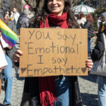 At the Women's March on March 8, 2025, from Washington Square Park to Union Square, a young woman stands smiling while holding a handmade cardboard sign that reads: "You say 'Emotional' I say 'Empathetic'" with small hearts decorating the corners. She is wearing a black long-sleeve top, blue jeans, and a deep red scarf wrapped around her neck. Her expression conveys confidence and resilience as she engages with fellow demonstrators.
To her left, another protester wearing a light pink T-shirt with a bold uterus design on the back and a black baseball cap appears to be engaged in conversation. The crowd in the background includes people holding various signs and LGBTQ+ pride flags, emphasizing the inclusive and intersectional nature of the event. A man wearing sunglasses and a pink bandana stands nearby, capturing the energy of the protest. The bright blue sky and leafless trees set the scene in an urban park filled with activism and solidarity.