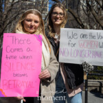 At the Women's March on March 8, 2025, in New York City, two women stand side by side holding impactful protest signs advocating for women’s rights and justice. The woman on the left wears a cream-colored coat over a soft pink top, accessorized with a pearl headband. She holds a bold pink sign with an inspirational quote that reads: "There comes a time when SILENCE becomes BETRAYAL", with the emphasized words written in a darker pink shade.
The woman on the right wears a long beige coat over a black outfit and black-framed glasses. She holds a white sign that reads: "We are the voices for WOMEN who NO LONGER HAVE ONE", with the words “WOMEN” and “NO LONGER HAVE ONE” highlighted in pink and purple. She wears leopard-print gloves, adding a touch of personal style to her protest attire.
The background features a crowd of demonstrators in Washington Square Park, many holding signs in solidarity, while leafless trees and city buildings frame the scene. The women’s expressions convey determination and resilience as they stand together in the movement.