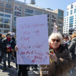 An elderly woman with short white hair and sunglasses participates in the Women's March on March 8, 2025, in Union Square, New York City. She holds a large handwritten protest sign on white poster board with red marker, reading:
"WOM STRONGER than any MAN
Musk is a Wacko!
$ NO MORE Amazon $
$ No more Tesla $
$ Money 4 U"
She wears a brown jacket with a red scarf and holds a bright yellow object in her left hand. Her determined expression and strong stance emphasize her activism against corporate billionaires. Surrounding her, a diverse group of protesters, many wearing pink hats and holding signs, fill the streets in solidarity for women's rights and economic justice. The background features Union Square with its iconic buildings and storefronts, capturing the energy of the event.
