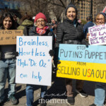Four demonstrators stand together at the Women's March on March 8, 2025, in Union Square, New York City, holding signs criticizing political corruption, authoritarianism, and foreign influence in U.S. politics. Each sign delivers a strong political message:
The first protester on the left holds a cardboard sign reading "DEFEND THE RULE OF LAW." They are wearing a black cropped jacket over a dark-colored top, light-wash jeans, and teal sneakers.
The second protester, an older woman in a red knit hat and red scarf, holds a white sign with blue handwritten text stating: "Brainless, Heartless. Maybe 'Dr. Oz' can help." She is wearing glasses, a black coat, and gloves.
The third protester, a woman in a black puffer jacket and beanie, holds a blue and yellow sign that says: "STOP PUTIN'S PUPPET FROM SELLING USA OUT."
The fourth protester on the right, wearing a gray beanie, glasses, and mustard-colored gloves, holds a white sign with red and blue text stating: "When we say EXECUTE the law, we don’t mean KILL IT!" She is also carrying a beige tote bag.
The crowd in the background consists of other activists holding banners and signs, engaging in discussions, and marching. Many are dressed in warm clothing, indicating a cold day. The setting is a lively, engaged demonstration for democracy and justice.