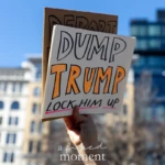 A demonstrator at the Women's March on March 8, 2025, raises a bold sign that reads "DUMP TRUMP - LOCK HIM UP" in large, hand-drawn letters, with "TRUMP" emphasized in orange. Behind it, a partially visible second sign appears to say "DEPORT." The protester’s hand is gripping both signs firmly, holding them high against the bright blue sky, with modern glass buildings and older architectural structures visible in the background.
The photo captures the charged political atmosphere of the march, where participants express their opposition to political figures and policies they believe threaten democracy, human rights, and women's rights. The Women's March, held annually, continues to be a powerful platform for advocacy on social justice issues.