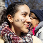 A young woman at the Women's March on March 8, 2025, displays the word "RESIST" boldly written in black ink across her right cheek. She smiles slightly, exuding confidence and determination. Her curly hair is pulled back, and she wears a small gold hoop earring. She is dressed warmly in a thick, multi-colored plaid scarf wrapped around her neck and a cream-colored coat, suggesting the chilly weather during the march.
In the background, other protesters can be seen, including an older woman wearing a navy blue beanie and glasses, dressed in a white winter coat. The setting is outdoors, with leafless trees visible, indicating the late winter or early spring season. The Women's March, taking place from Washington Square Park to Union Square in New York City, is a powerful gathering advocating for women's rights, equality, and social justice.