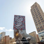 A protester at the Women's March on March 8, 2025, in Union Square, NYC, raises a colorful sign reading "My Body, My Choice" in bold letters wrapped in a ribbon design. The background of the sign is filled with a vibrant floral pattern, symbolizing empowerment and reproductive rights. The message, a rallying cry for bodily autonomy, stands out against the clear blue sky as it is held high among a crowd of demonstrators.
The low-angle shot captures the urban backdrop of New York City, featuring tall buildings and streetlights, reinforcing the significance of the protest in a major metropolitan setting. The march is part of a larger movement advocating for women's rights, reproductive freedom, and gender equality.