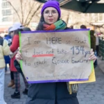 A protester at the Women's March on March 8, 2025, in Union Square, NYC, stands in front of a subway entrance holding a handmade sign that reads: "I am HERE so that it does not take 134 yrs to reach GENDER EQUALITY." The sign, crafted on brown cardboard, is highlighted with red and blue underlines, emphasizing urgency and frustration over the slow pace of progress toward gender equality.
The protester wears a purple beanie, clear glasses, a dark blue puffer coat, and a multicolored scarf with shades of red, green, yellow, and orange. The layered winter attire suggests a chilly day, but their firm grip on the sign and determined expression display unwavering commitment to the cause. The urban setting of Union Square, with other demonstrators visible in the background, highlights the large turnout and collective advocacy for women's rights.