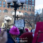 A crowd of demonstrators fills Union Square, NYC, during the Women's March on March 8, 2025, holding up various signs advocating for women's rights and gender equality. In the center of the image, a sign reads: "I AM LIFE, I AM STRENGTH, I AM WOMAN," a powerful declaration of resilience and empowerment. Many participants wear pink knitted hats, a symbol of feminist solidarity, while others hold banners and placards with messages addressing political and social issues.
The backdrop features tall city buildings and streetlights, emphasizing the urban setting. Protesters are seen actively engaging in the demonstration, raising signs and marching together in unity. The diversity of messages and the visible passion in the crowd showcase the strength and determination of those advocating for change.
