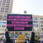 At the Women's March 2025 in Union Square, NYC, a protester raises a striking black and pink sign displaying a controversial quote:
"When you're a star, they'll let you do it... Grab them by the pussy."
The protester, dressed in a black coat, firmly holds the sign above their head, ensuring its visibility among the crowd of demonstrators. Many other participants are in the background, some wearing pink hats, a reference to past feminist movements like the Women's March Pussyhat Project. The location, Union Square, NYC, is filled with protesters carrying various signs advocating for women's rights, equality, and resistance against misogyny.
The quote on the sign is a direct reference to Donald Trump's infamous 2005 remarks, often cited as a symbol of sexism and the abuse of power. By displaying the statement in a bold, attention-grabbing format, the protester is likely condemning misogyny and raising awareness about sexual misconduct in politics and society. The march brings together activists, feminists, and allies, using powerful imagery and words to demand accountability and advocate for women's empowerment.