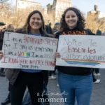 Two women stand side by side, smiling as they proudly hold signs with feminist messages at the Women's March 2025 in Union Square, NYC. The woman on the left holds a sign featuring a quote from jazz singer Dianne Reeves' song "Endangered Species", which reads:
"I am an ENDANGERED SPECIES but I sing no victim’s song.
I am a WOMAN, I am an ARTIST.
I KNOW WHERE MY VOICE BELONGS."
— Dianne Reeves
She is dressed in a black jacket over a blue top and has long dark hair. Her handwritten sign is decorated with red, black, and blue lettering, emphasizing key words for empowerment.
The woman on the right holds a sign with the famous Latin phrase:
"Nolite te bastardes carborundorum"
which translates to "Don’t let the bastards grind you down", a phrase popularized by Margaret Atwood’s novel The Handmaid’s Tale. Her sign is decorated with multicolored lines at the bottom. She is wearing a black hoodie with orange text across the front, has curly brown hair, and her broad smile conveys confidence and determination.
The background captures the energy of the march, with a crowd of activists holding signs, engaging in conversations, and standing in solidarity for women’s rights, gender equality, and social justice. The setting is Union Square, NYC, with the park's familiar architecture and trees visible, along with attendees wearing winter clothing, suggesting a cold but sunny March day.