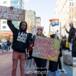 A powerful moment at the Women's March 2025 in Union Square, NYC, where two protesters stand side by side holding signs advocating for protection and transgender rights. The man on the left, wearing glasses, a black hoodie with the words “I AM BLACK HISTORY”, and red pants, raises a cardboard sign reading "REAL MEN PROTECT" in bold white letters. His stance and expression exude strength and conviction.
Next to him, a woman with a determined expression wears a decorative headpiece resembling a crown, a black coat, and black pants while holding a bright yellow and pink spray-painted sign that reads: "DEFEND TRANS SISTERS". In her left hand, she clutches a red megaphone, symbolizing the vocal activism present in the march.
The background captures the broader scene of the march, with diverse protesters waving pride flags, holding signs, and taking photos. The setting is filled with energy, with banners promoting LGBTQ+ rights, gender equality, and social justice. The "LOVE" pride flag is prominently visible in the crowd, reinforcing the spirit of inclusivity and solidarity. The street is painted with colorful artwork, adding vibrancy to the protest scene.