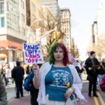 A passionate protester marches through Union Square, NYC, during the Women's March 2025, holding a handmade sign reading "PAWS OFF LAWS OFF" in bold purple and blue letters, decorated with flowers and paw prints. She wears a striking ensemble featuring a blue graphic t-shirt with "The Female Gaze: Petrify the Patriarchy" printed across the front, referencing Medusa as a feminist symbol. Over her shirt, she wears a flowy, white skirt and a light cardigan, adding an ethereal touch to her outfit.
Her vibrant two-toned curly hair, dyed green on one side and pink on the other, stands out, reflecting the spirit of self-expression and empowerment. She holds a yellow flower in one hand, a subtle yet powerful symbol of resistance and hope. The background shows a bustling protest scene with other demonstrators holding signs, police presence, and towering city buildings lining the march route.
This image captures the fierce advocacy for women's autonomy, highlighting themes of bodily rights, legal justice, and feminist solidarity during one of NYC's most significant annual marches.