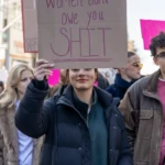 A determined protester at the Women's March 2025 in New York City raises a cardboard sign with a powerful statement: "Women don't owe you SHIT", written in bold, capitalized letters. She wears a black puffer jacket over a dark green hoodie, exuding a strong and confident presence as she marches with fellow activists. Her expression is one of quiet defiance, embodying the spirit of the movement.
To her right, a man wearing a brown jacket over a pink sweater walks alongside her, looking towards the camera. Behind her, other marchers hold signs with feminist messages, including one with a bright pink background. The bustling cityscape and streetlights frame the scene, emphasizing the urban setting of this mass mobilization for gender equality and women's rights.
This image captures the unwavering stance of modern feminism, with demonstrators demanding respect, autonomy, and systemic change in a world where women's voices continue to fight for recognition.