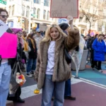 A young woman stands confidently at the Women's March 2025 in New York City, holding up a handmade sign that resembles a TIME magazine cover. The sign reads "Person of the Year TIME – Gisele Pelicci", featuring a simple sketch of a woman with glasses. She wears a luxurious faux fur coat over a white T-shirt with a feminist message, paired with blue denim jeans and black shoes. Her blonde hair is styled in loose waves, and she gazes directly at the camera with a composed and determined expression.
In her left hand, she holds a small yellow flower, symbolizing hope and resilience. To her right, another protester holds a pink sign with an assertive feminist statement, while behind her, a diverse crowd marches with various placards and banners advocating for women's rights and social justice. The setting is Union Square, with the city's tall buildings and the bustling energy of the demonstration serving as the backdrop.
This image encapsulates the spirit of empowerment and recognition of women’s contributions, as marchers celebrate figures they admire and push for systemic change.