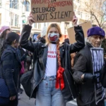 At the Women's March 2025 in New York City, a protester holds a handmade cardboard sign that reads:
"COUNTLESS BANS ON WOMEN'S MARCHES IN KAZAKHSTAN. SILENCED, NOT SILENT."
The protester, a young woman with long red hair in a braid tied with a bright red ribbon, is wearing a black leather jacket, a white t-shirt with "Smash the Patriarchy" written on it, and light-washed denim jeans. She also wears a face mask featuring the design of the Kazakhstan flag, symbolizing solidarity with activists in the country. Her stance is strong as she raises her sign high above her head.
To her right, an older woman wearing a purple beret and a dark coat holds another protest sign, which reads "NO! To fascism", indicating the broad political concerns present at the march. The background is filled with other demonstrators, many carrying signs, dressed in layers for the chilly weather, and walking along Union Square as part of the rally. The urban setting of New York City’s Union Square is prominent, with tall buildings and storefronts lining the streets.
This image captures the international scope of the Women's March, showing solidarity for women's rights beyond the U.S. and drawing attention to issues of gender equality and protest suppression worldwide.