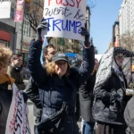 At the Women's March 2025 in New York City, a determined protester raises a bold sign reading:
"My PUSSY won't be TRUMPED" in a mix of black, red, and blue lettering.
The woman, standing strong in the middle of the street, is wearing a dark blue puffer coat, a black baseball cap with white embroidered text, gloves, and a black crossbody bag. Her serious expression conveys resolve as she participates in the march alongside a crowd of fellow demonstrators.
To her left, another protester in a gray coat and orange scarf holds a separate sign, though the text is partially obscured. To her right, a person dressed in a striking black-and-white polka-dot veil, black gloves, a lace-trimmed jacket, and vibrant cartoon-patterned leggings stands in the crowd, adding to the diversity of voices and styles present at the event.
The backdrop features tall buildings and New York City streets filled with demonstrators, signs, and activism, emphasizing the march's scale and significance. The protest calls attention to women's rights, political resistance, and bodily autonomy, key themes of the Women's March movement.