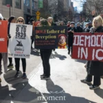 At the Women's March 2025 in New York City, a group of protesters stands in the middle of the street holding striking signs that criticize Elon Musk, authoritarianism, and the state of democracy.
The protester on the right, wearing a black coat and black pants, holds a large red banner with white painted letters that reads:
"DEMOCRACY IN ICU", implying that democracy is in a critical state.
To her left, other demonstrators hold signs with text targeting Elon Musk and his influence. One sign, painted red, lists words vertically, spelling out "ELON MUSK" with descriptors such as "Exploitative, Ludicrous, Obsessive, Nazi," "Megalomaniac, Unhinged, Satanic, Kleptocrat." Another sign reads, "Goes from 0 to 1939 in 3 seconds" with an image of a Tesla and the phrase "Tesla The Swasticar", drawing a historical parallel between Musk's actions and authoritarian regimes.
Another protester holds a sign resembling a movie poster, reading:
"DOGEfather of Deception – In theaters now starring #ElonMania", a play on Musk's involvement in cryptocurrency and his media presence.
The street is crowded with marchers, while NYPD officers stand nearby monitoring the demonstration. In the background, food trucks and buildings line the street, adding an urban atmosphere to the protest scene. The clear sky and bright sunlight illuminate the moment, highlighting the intensity and passion of the demonstrators.