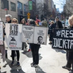 During the Women's March 2025 in New York City, demonstrators held up bold and satirical signs targeting Donald Trump, Elon Musk, and authoritarianism.
On the left, a protester holds a large notebook-style sign that mocks Trump’s speech patterns with exaggerated quotes, titled:
"Welcome to 1st grade, Trumpy! Time to Make Recess Great Again."
At the bottom, it is graded with a red "F", ridiculing Trump's rhetoric and policies.
Next to this sign, another protester holds a black-and-white poster that reads:
"Goes from 0 to 1939 in 3 seconds" with an image of a Tesla and the phrase "Tesla The Swasticar." This sign equates Elon Musk’s influence with historical authoritarian regimes.
To the right, a large open book-style sign reads:
"A Tale of a Statesman and a Conman," suggesting a comparison between two historical leaders—one praised by history, the other facing legal troubles.
On the far right, a protester in a black coat and crocs holds a striking black-and-white sign that reads:
"#RESIST DICK-TRAITOR", a direct jab at Trump’s leadership and influence.
The march takes place on a busy NYC street, with people walking and NYPD officers monitoring the protest. The sun casts sharp shadows, highlighting the intensity of the moment. Other demonstrators and urban buildings serve as a backdrop, reinforcing the grassroots resistance energy of the event.
