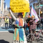 A demonstrator at the Women's March 2025 in Union Square, NYC, holds a large yellow sign with a powerful message:
"IF WOMEN WERE SAFE, THERE WOULDN’T BE A WAR WAGED ON THEIR BODIES."
Dressed in a striking, multicolored geometric-patterned coat, a matching knit scarf, and a colorful bucket hat, the protester stands with arms raised, amplifying her message about women’s safety and bodily autonomy.
Behind her, a crowd of marchers waves pride flags, and a participant in an orange beanie films the protest on their phone. A bicycle leans against a street pole, adding to the dynamic urban setting. The street signs for Union Square West and East 14th Street place the demonstration firmly in the heart of Manhattan.
The clear blue sky and bright sunlight create sharp contrasts, making the protest's energy and urgency even more striking. Other marchers and activists fill the street, reinforcing the spirit of collective resistance.
